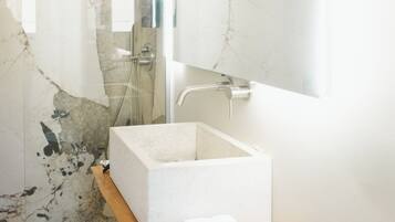 A bathroom with a glass shower enclosure, a marble-tiled wall, a white sink, and a wooden shelf with towels.