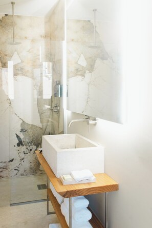 Un baño con cabina de ducha de vidrio, pared revestida de mármol, lavamanos blanco y repisa de madera con toallas.