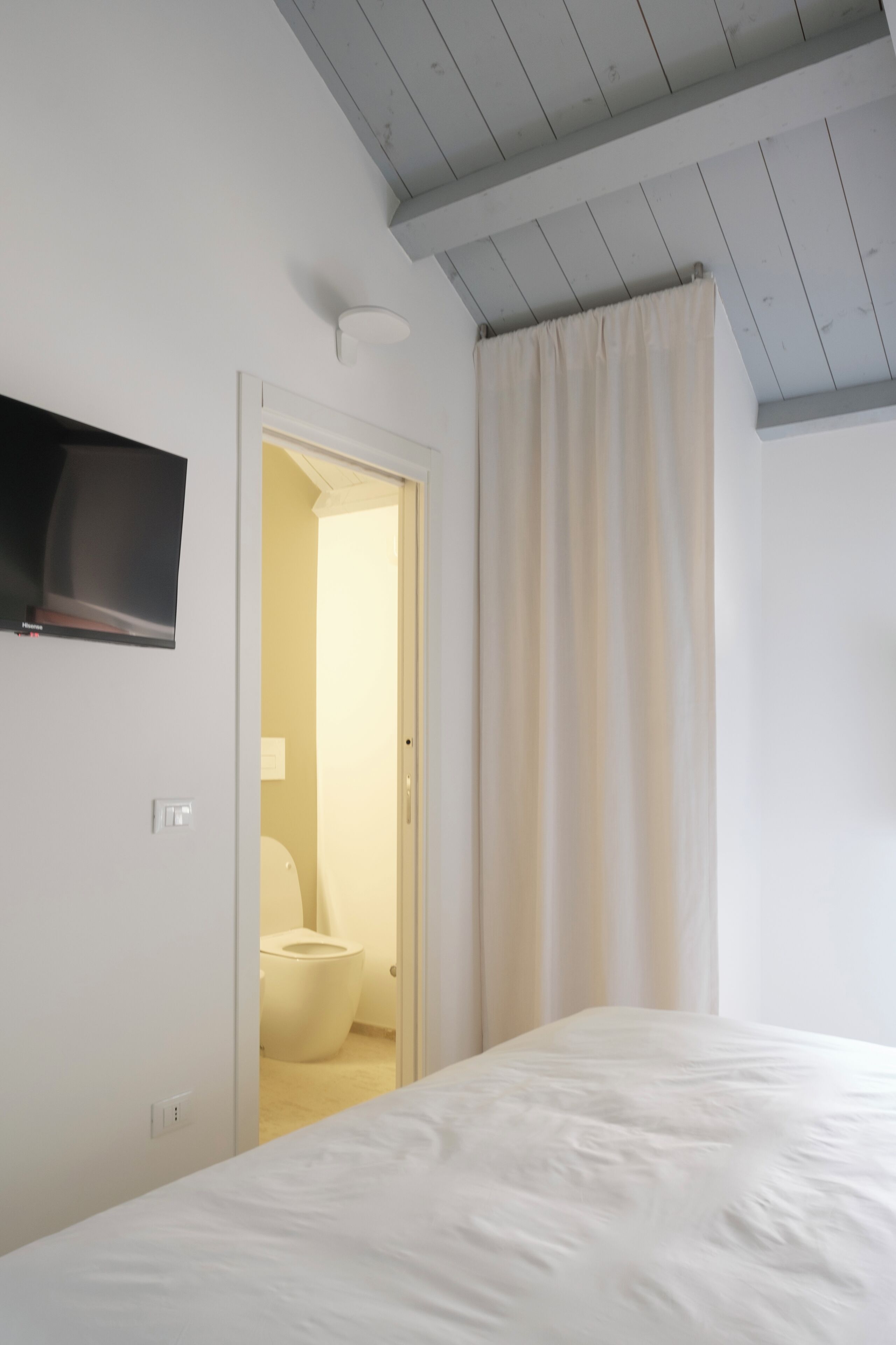 Une chambre à coucher avec un lit, un téléviseur fixé au mur et une salle de bain visible à travers une porte ouverte.