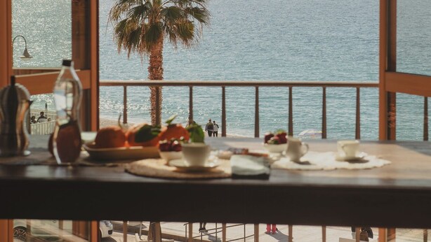 A table with dishes and a bottle, overlooking a body of water with a palm tree and distant mountains. - La Balza di Scilla (Scilla)