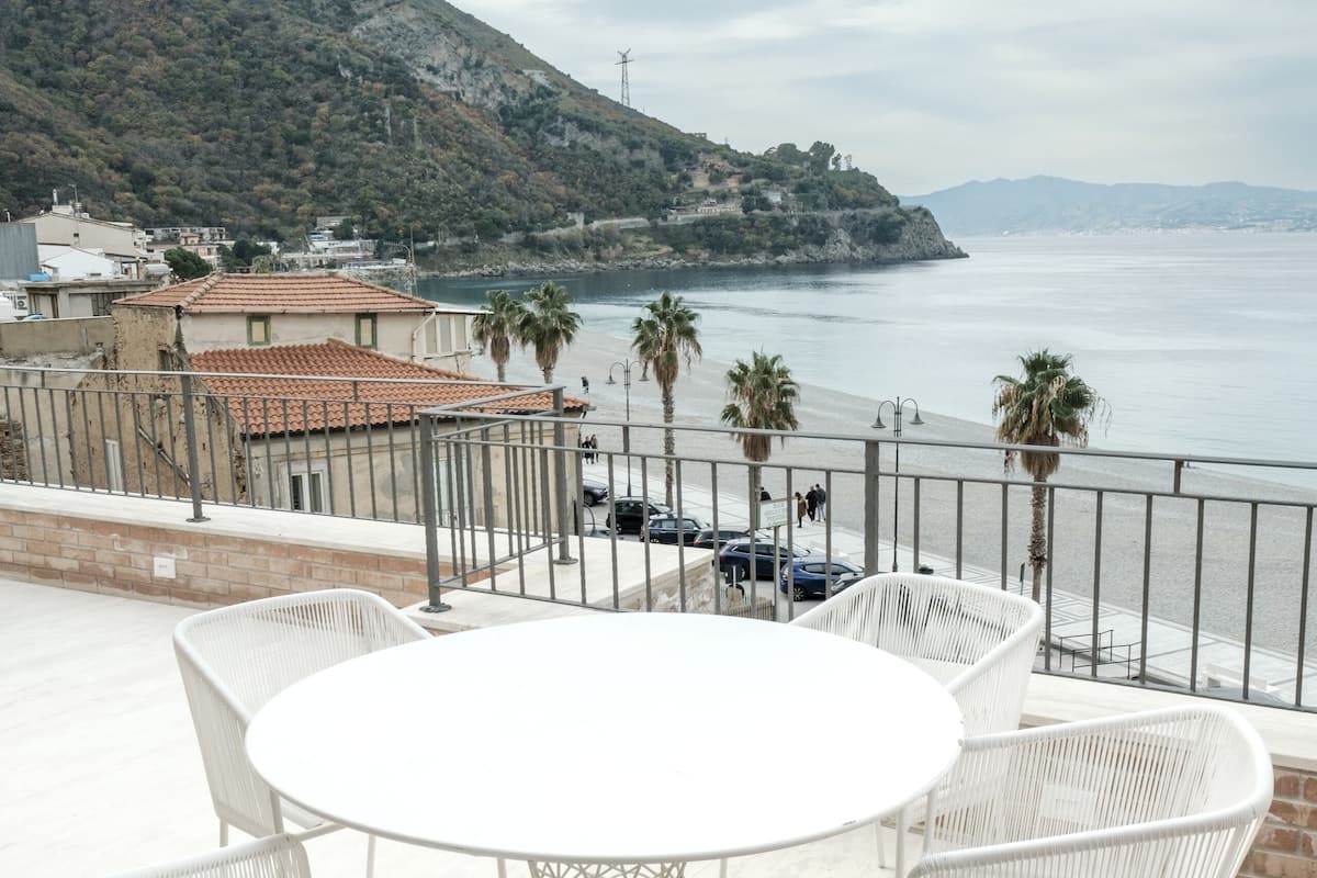 A terrace with a white round table and chairs, overlooking a beach and a mountainous landscape.