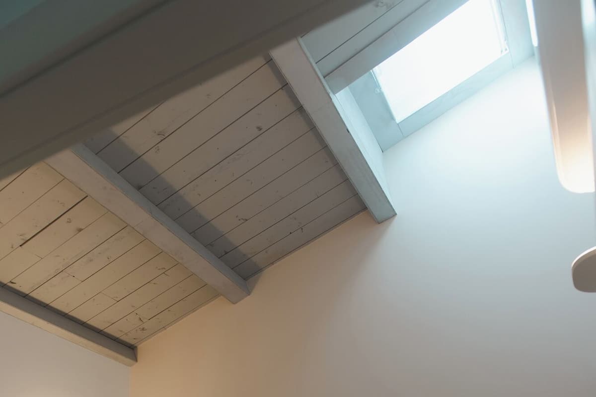 A bedroom with a bed, pillows, a nightstand, a wall-mounted lamp, and a skylight.