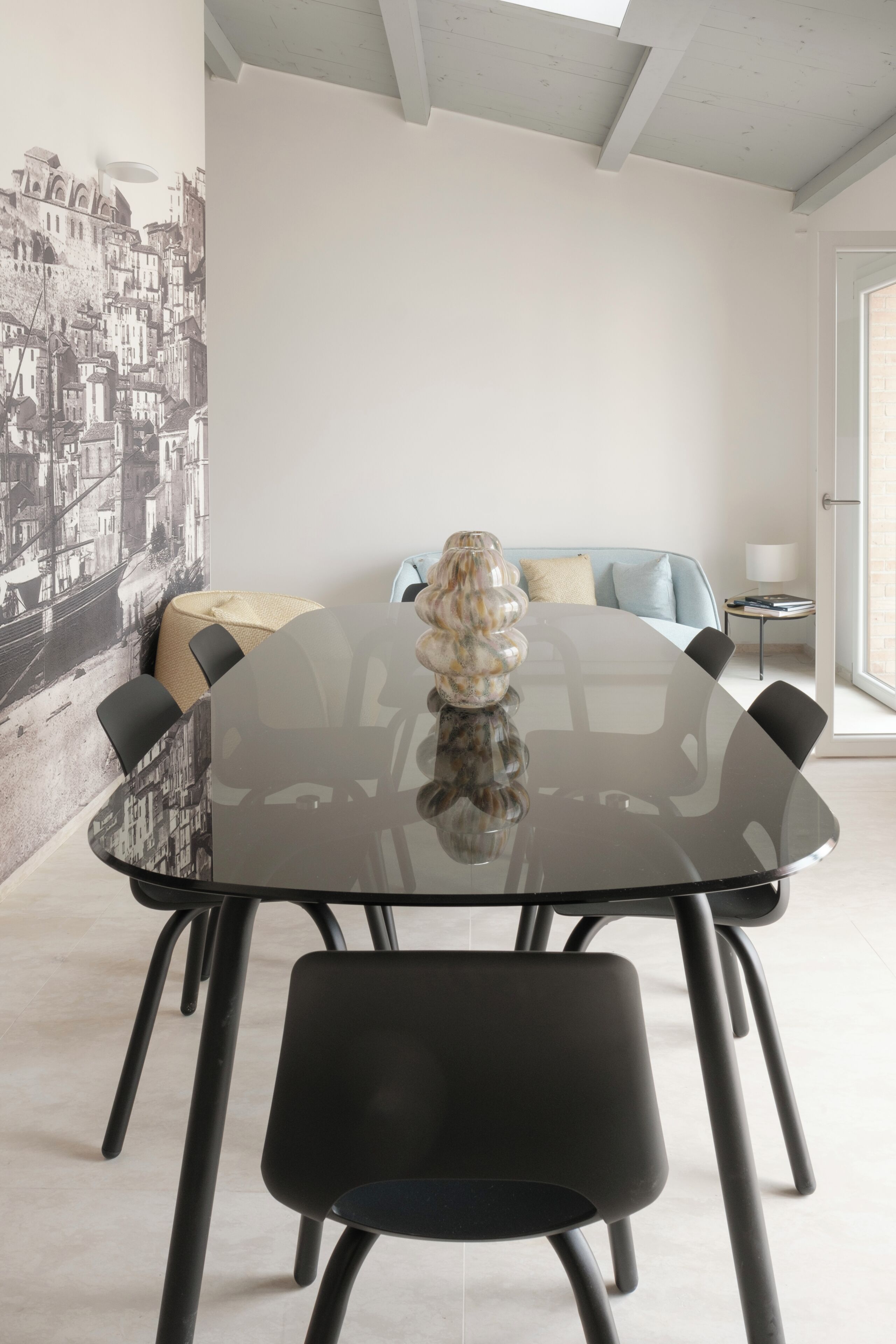 A modern dining area with a glass table, black chairs, a decorative vase, a sofa, and a cityscape mural on the wall.