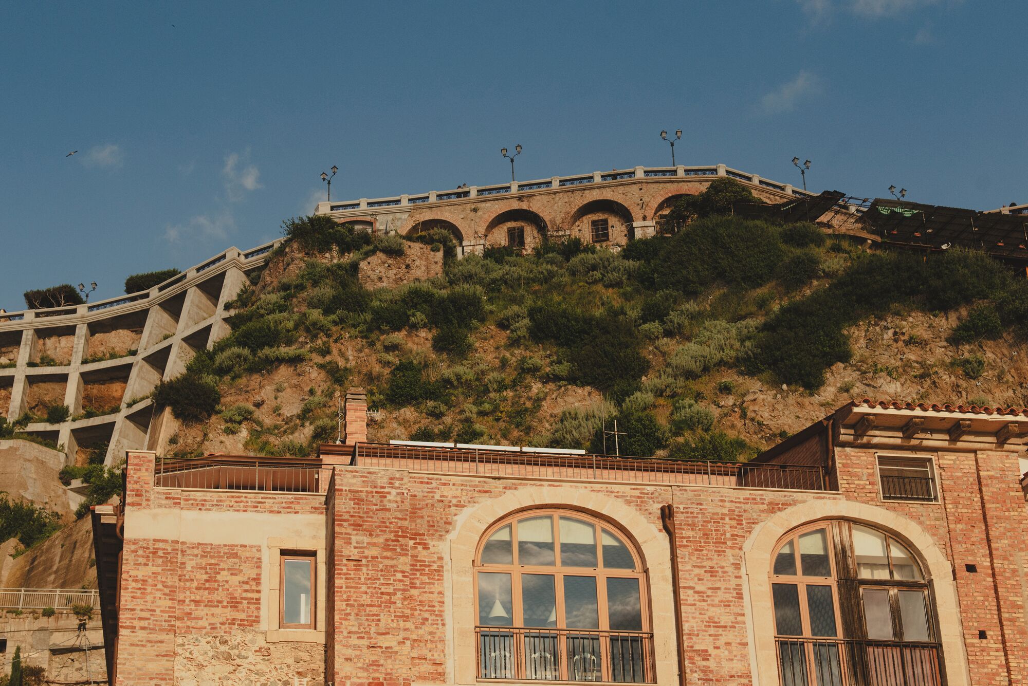 Ein Gebäude mit Bogenfenstern und einem Balkon, auf einem Hügel gelegen, mit einer Brücke im Hintergrund.