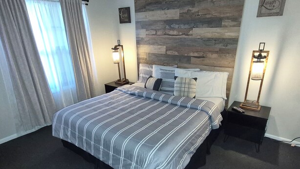 Quarto de hotel com cama, duas mesas de cabeceira, luminária na parede, janela com cortinas e parede com textura de pedra.