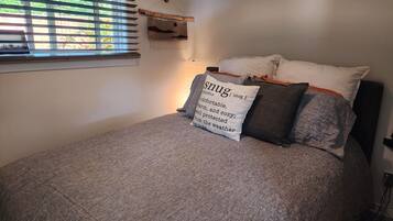 Une chambre à coucher avec un lit, une fenêtre avec des stores, une lampe de chevet et une étagère fixée au mur.
