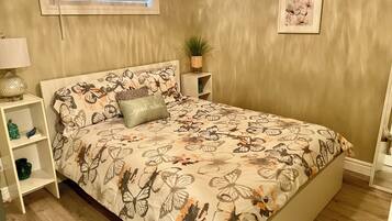 Een slaapkamer met een bed, nachtkastje, plank, raam met jaloezieën en een ingelijst schilderij aan de muur.