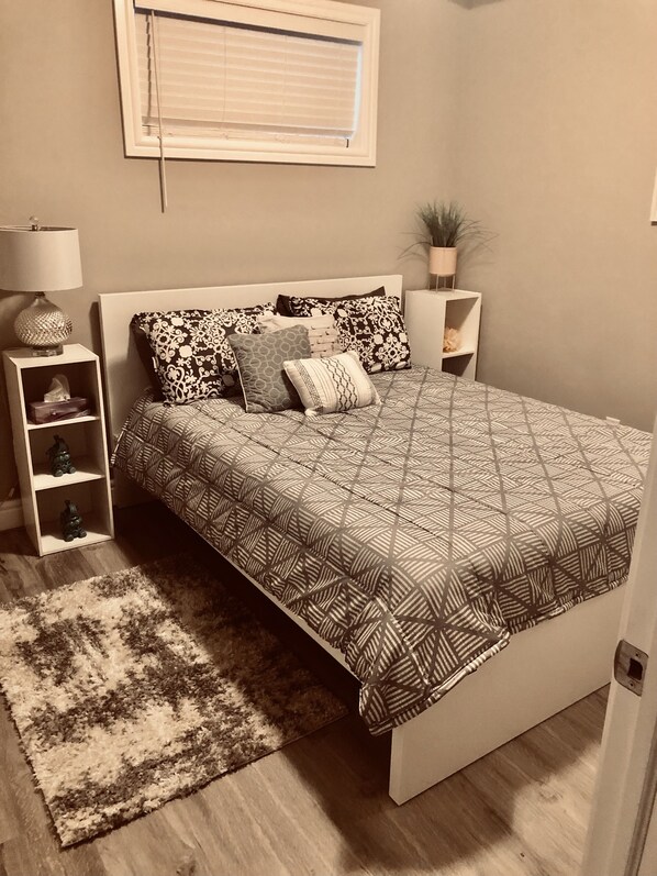 A bedroom with a bed, a bedside table, a lamp, a plant, and a window with blinds.