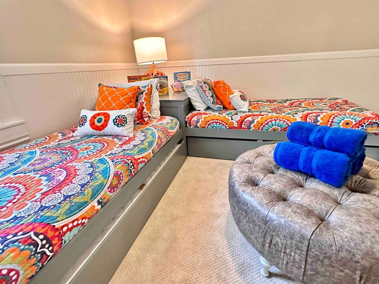 A hotel room with two beds, each with colorful patterned bedding and multiple pillows. There is a tufted ottoman with blue towels on it.