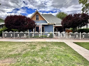 A two-story house with a blue exterior and a gray roof, surrounded by a white fence with decorative elements. - Pleasant Downtown Haven *0.3 Miles *Pet Friendly (Prescott)