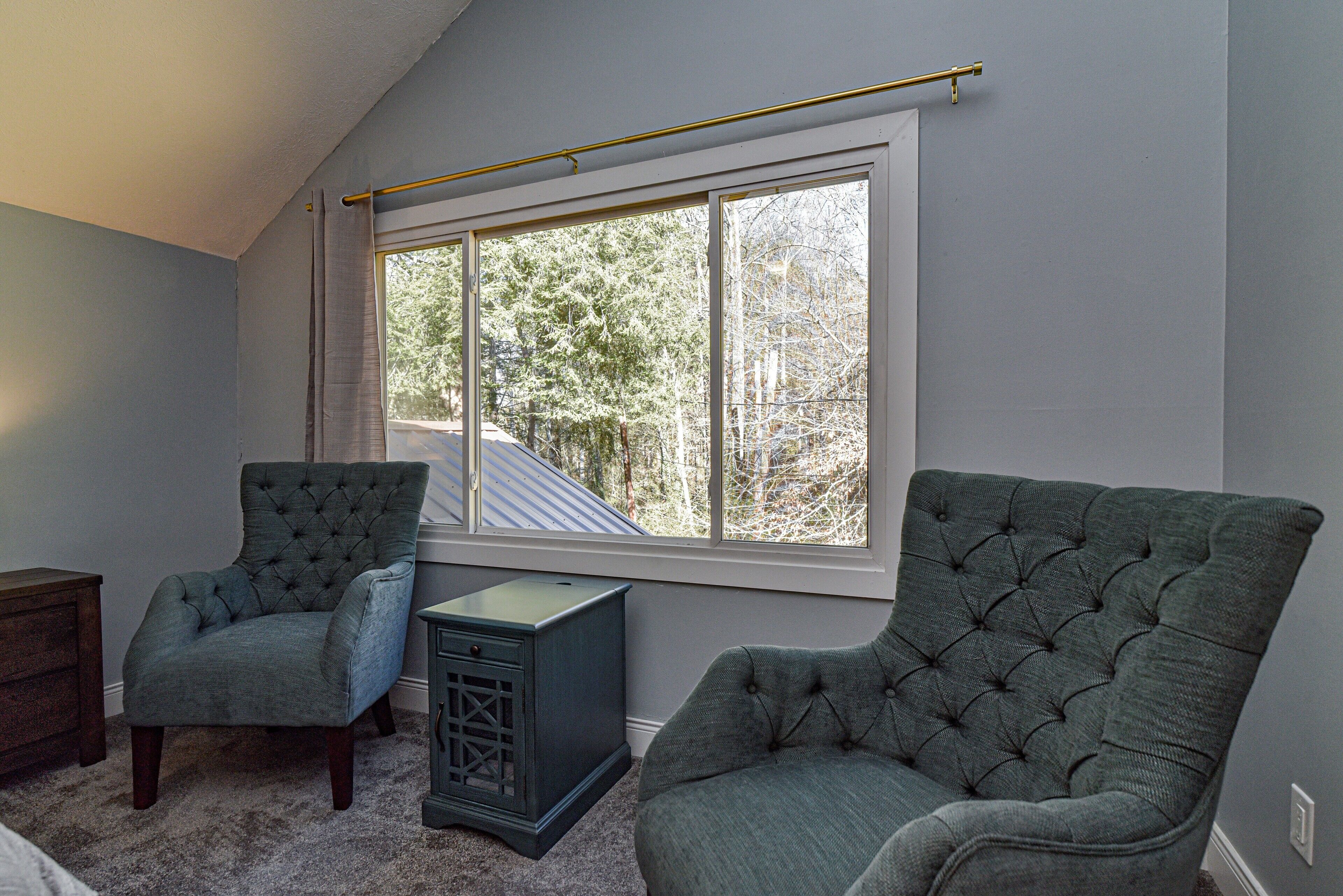 A room with a large window, two upholstered chairs, a small cabinet, and a carpeted floor.