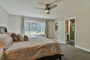 A bedroom with a large bed, a ceiling fan, a window with curtains, and a door leading to a bathroom. - Kingdom on Knollwood (Asheville)