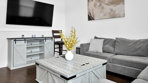 Una sala de estar moderna con un televisor de pantalla plana, un sofá gris, una mesa de centro con un jarrón blanco y un armario de estilo rústico corredizo.