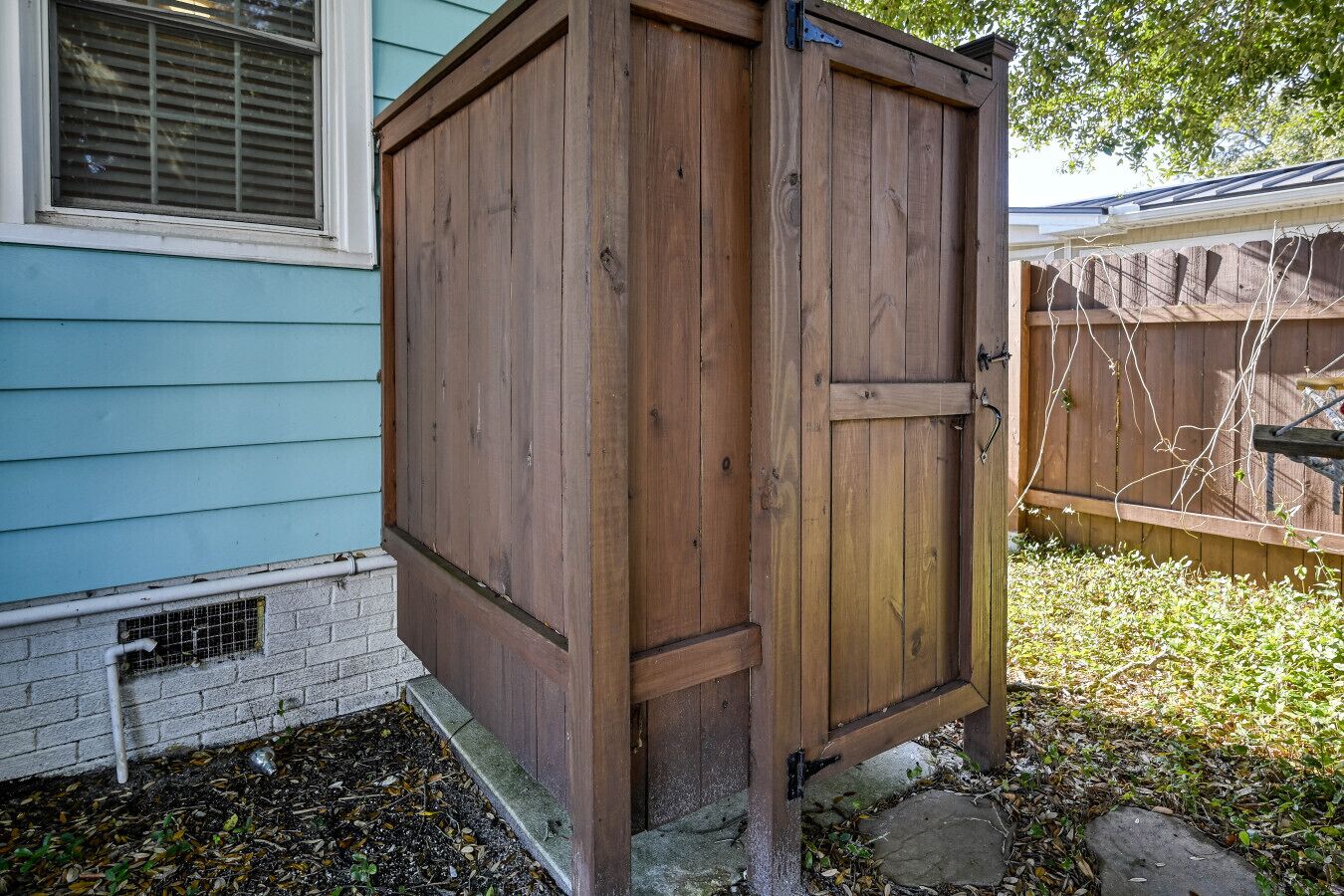 A wooden shed with a door, positioned next to a house with light blue siding and a window with blinds.