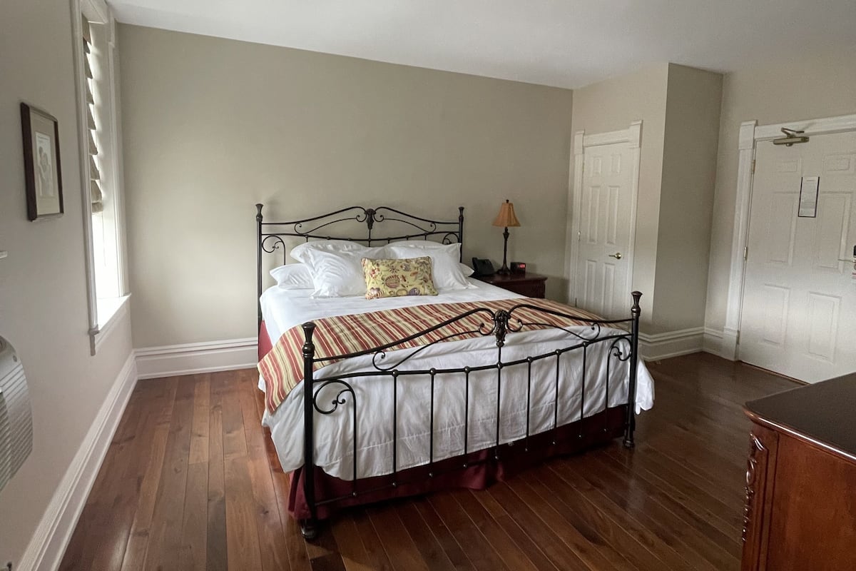 A bedroom with a wooden floor, a metal bed frame, a bedside table with a lamp, and a door.