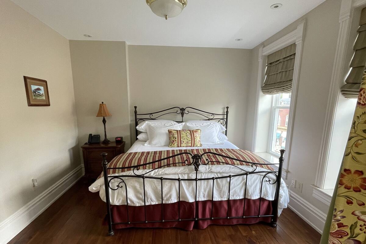 A bedroom with a wooden floor, a metal bed frame, a bedside table with a lamp, and a window with curtains.