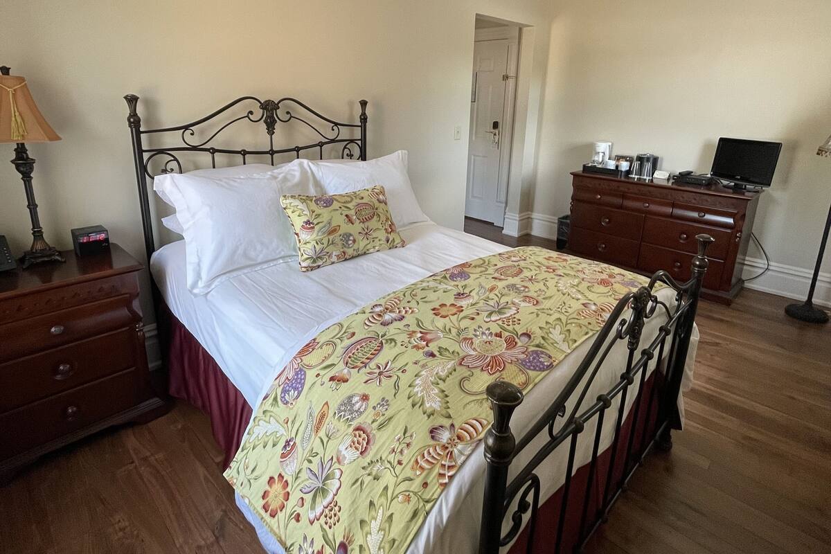 A neatly made bed with a floral bedspread, a wooden nightstand with a lamp, and a dresser with a flat-screen TV.