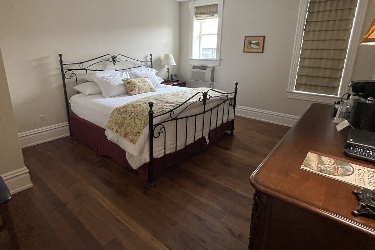 A bedroom with a wooden floor, a metal bed frame, a bedside table, and a window with blinds.
