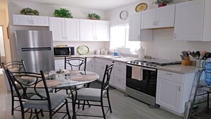 A modern kitchen with white cabinets, a stainless steel refrigerator, a microwave, and a gas stove. There is a dining table with chairs and a small potted plant on the counter.