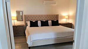 A neatly made bed with a tufted headboard, two bedside tables with lamps, a mirror, and a wall-mounted air conditioner.