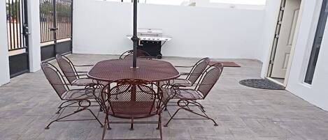 Une terrasse avec une table ronde et des chaises, un barbecue et un parasol rouge.
