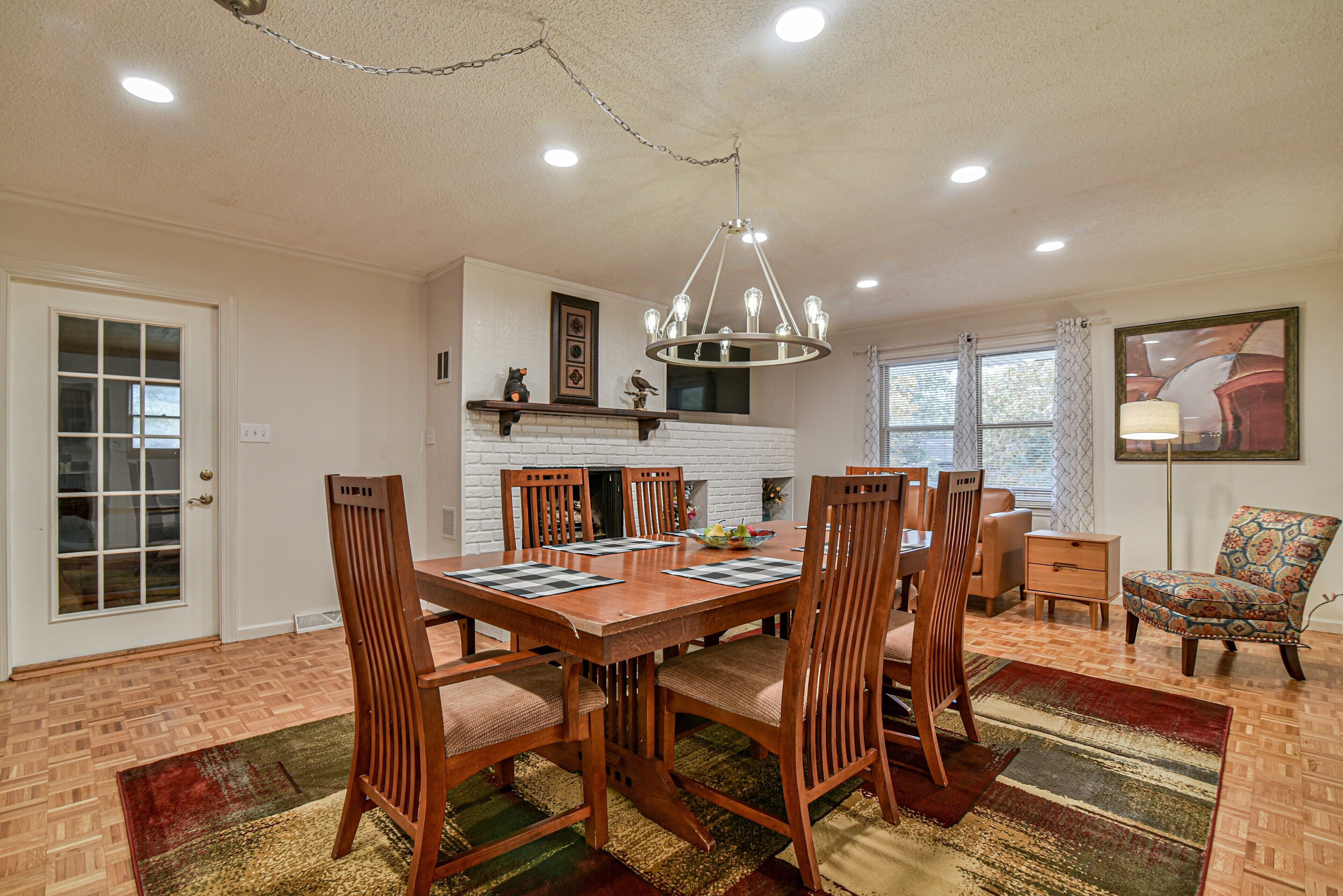 A dining room with a wooden table, chairs, and a chandelier. There is a fireplace, a window with curtains, and a patterned rug.