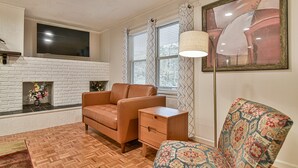 A living room with a white brick fireplace, a brown leather sofa, a patterned armchair, a small wooden nightstand, and a floor lamp. - Forest Run - 4 Bedrooms with hot tub (Asheville)
