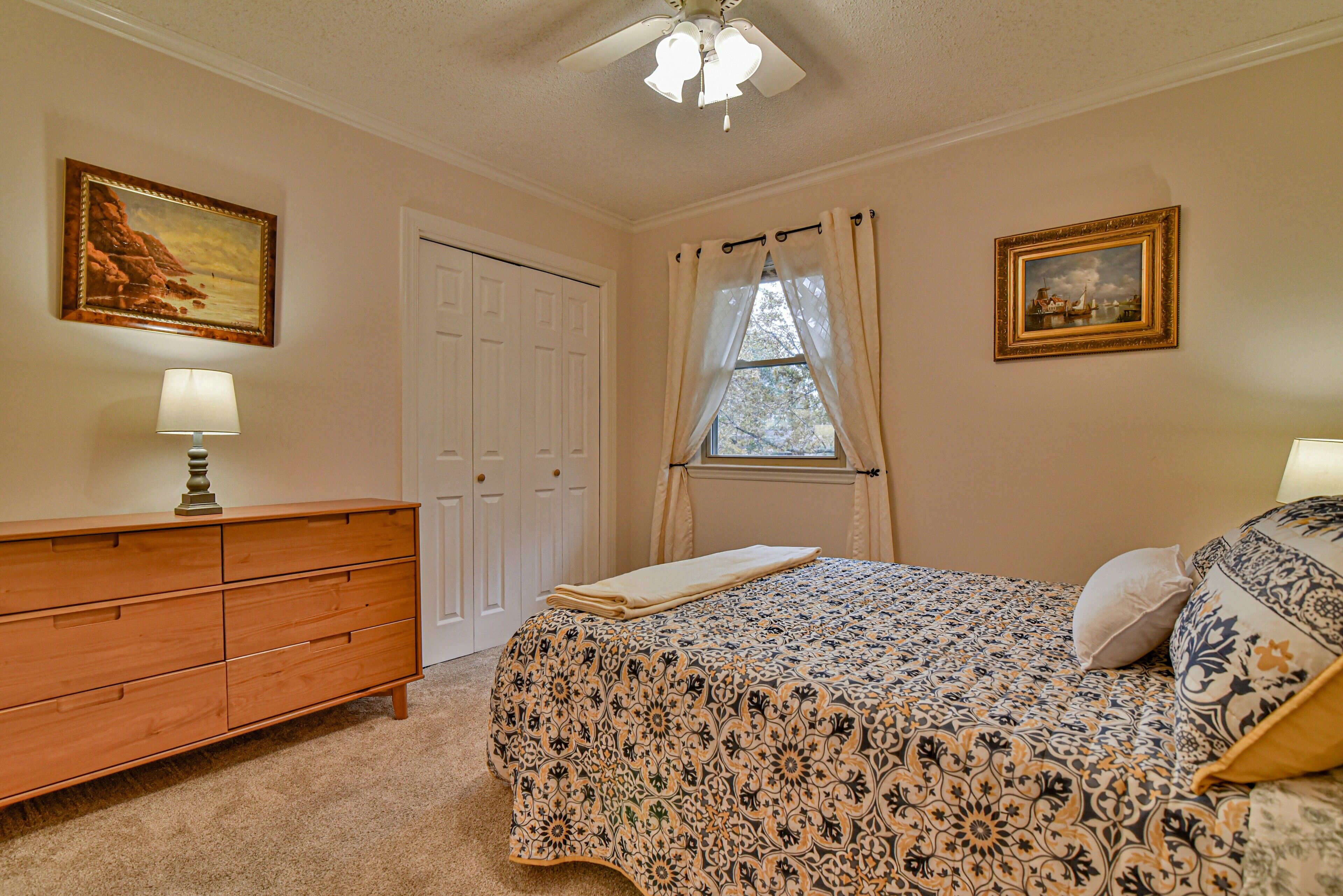 A bedroom with a bed, a dresser, a window with curtains, and two framed pictures on the wall.
