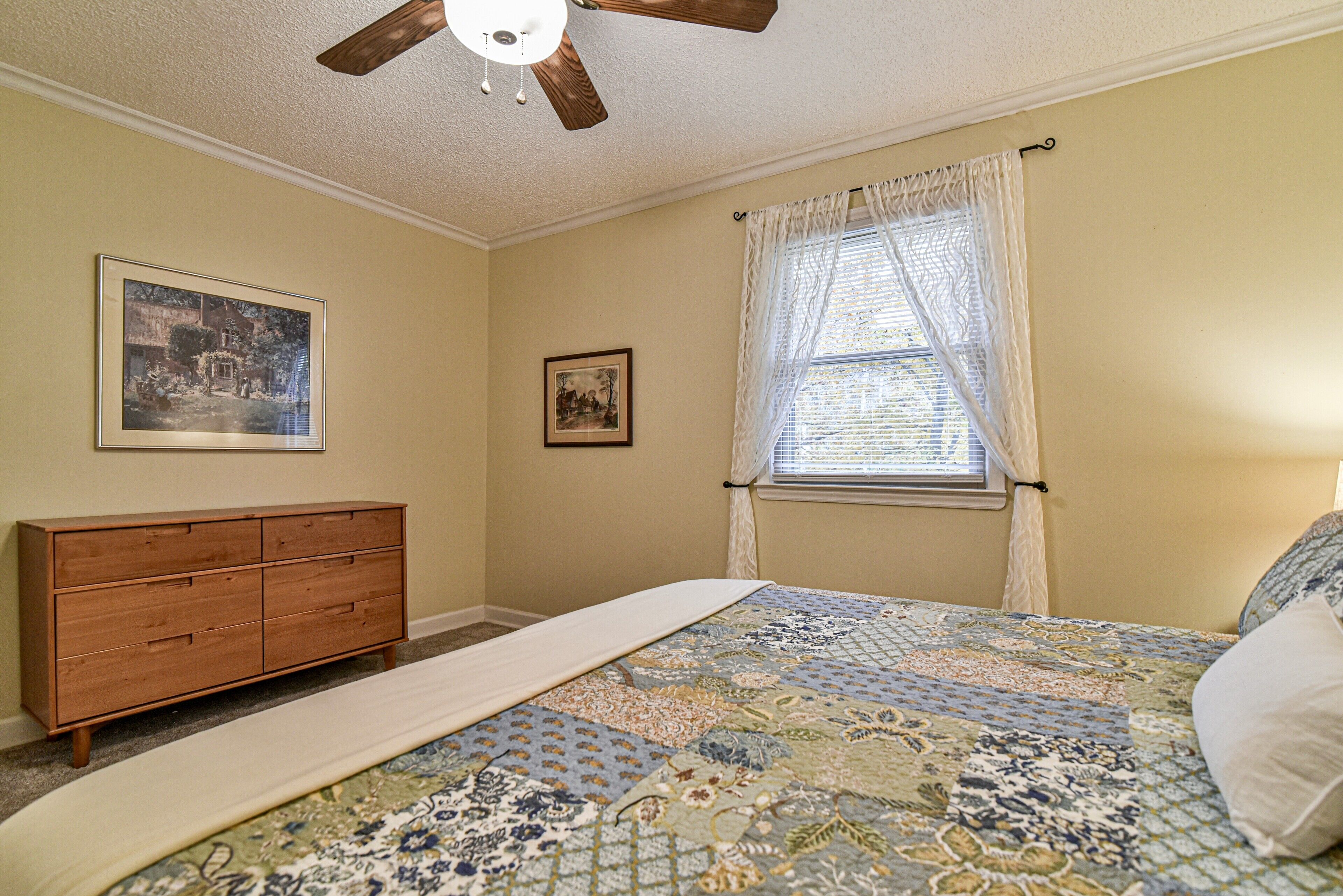 Un dormitorio con cama, cómoda de madera, dos cuadros en la pared, ventana con cortinas y ventilador de techo.