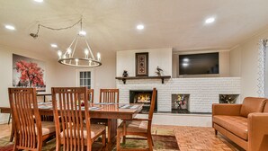A dining area with wooden chairs and a table, a fireplace with a white brick surround, and a television mounted above it. - Forest Run - 4 Bedrooms with hot tub (Asheville)