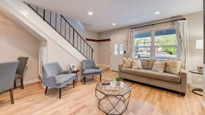 A living room with a staircase, a sofa, two armchairs, a coffee table, and a window with curtains.