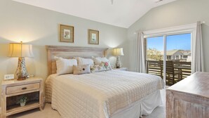 A bedroom with a large bed, two bedside tables, a nightstand, a wooden headboard, and a view of the outside through a sliding door. - Charming 3-Bed Townhome Steps from the Beach (Kill Devil Hills)