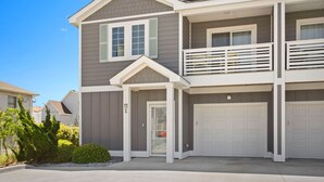 A two-story residential building with a garage, a balcony on the upper floor, and a small front yard. - Charming 3-Bed Townhome Steps from the Beach (Kill Devil Hills)