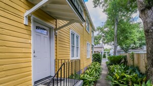 A yellow house with a white door and windows, a front porch with a black railing, and a pathway lined with green plants.