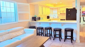 A modern kitchen with a central island, bar stools, and a white sofa in a bright room with large windows.