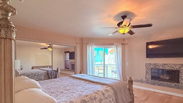 A bedroom with a large bed, a ceiling fan, a fireplace, and a TV mounted on the wall.