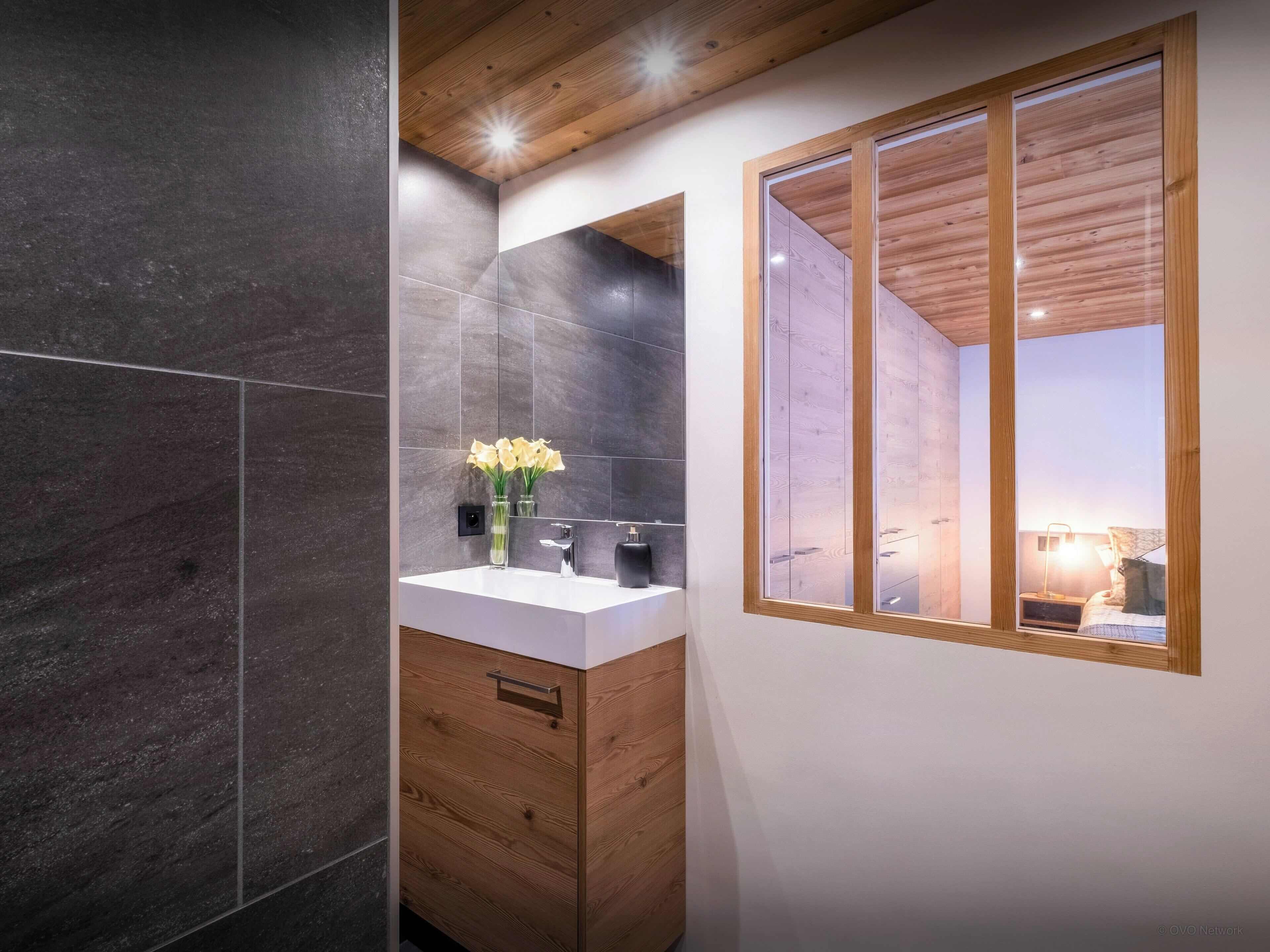 Un bagno moderno con piastrelle scure, un mobile lavabo in legno, uno specchio e una finestra con cornice in legno.