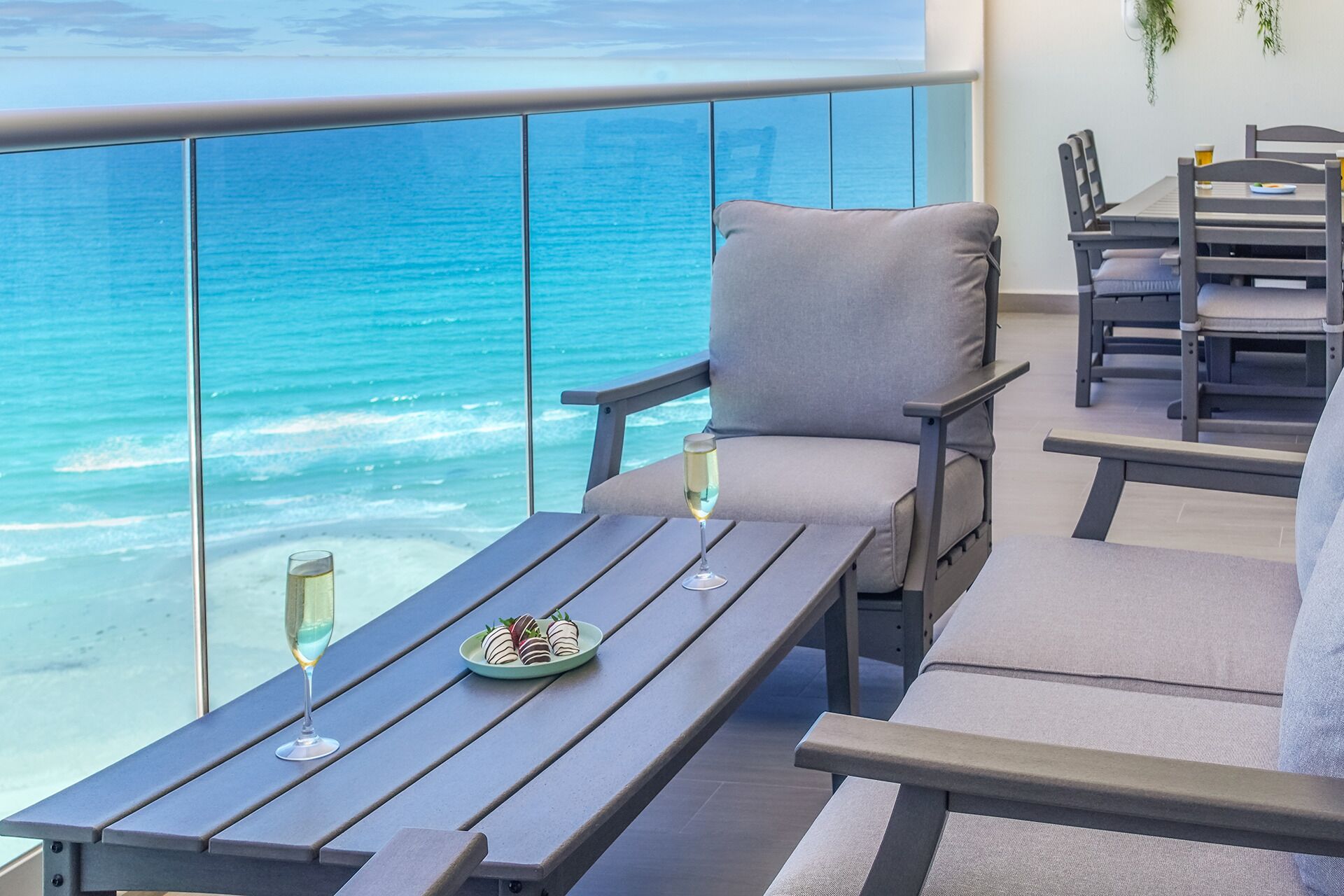 A balcony with a view of the ocean, a table with a plate of food and a glass, and a chair with a cushion.