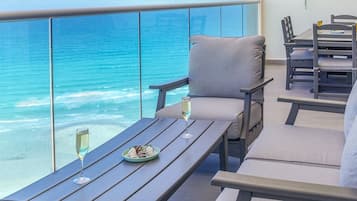 A balcony with a view of the ocean, a table with a plate of food and a glass, and a chair with a cushion.