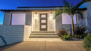 Ein modernes Haus mit gut beleuchtetem Eingang, einer Garagentür und einem kleinen Garten mit Pflanzen und einer Palme.