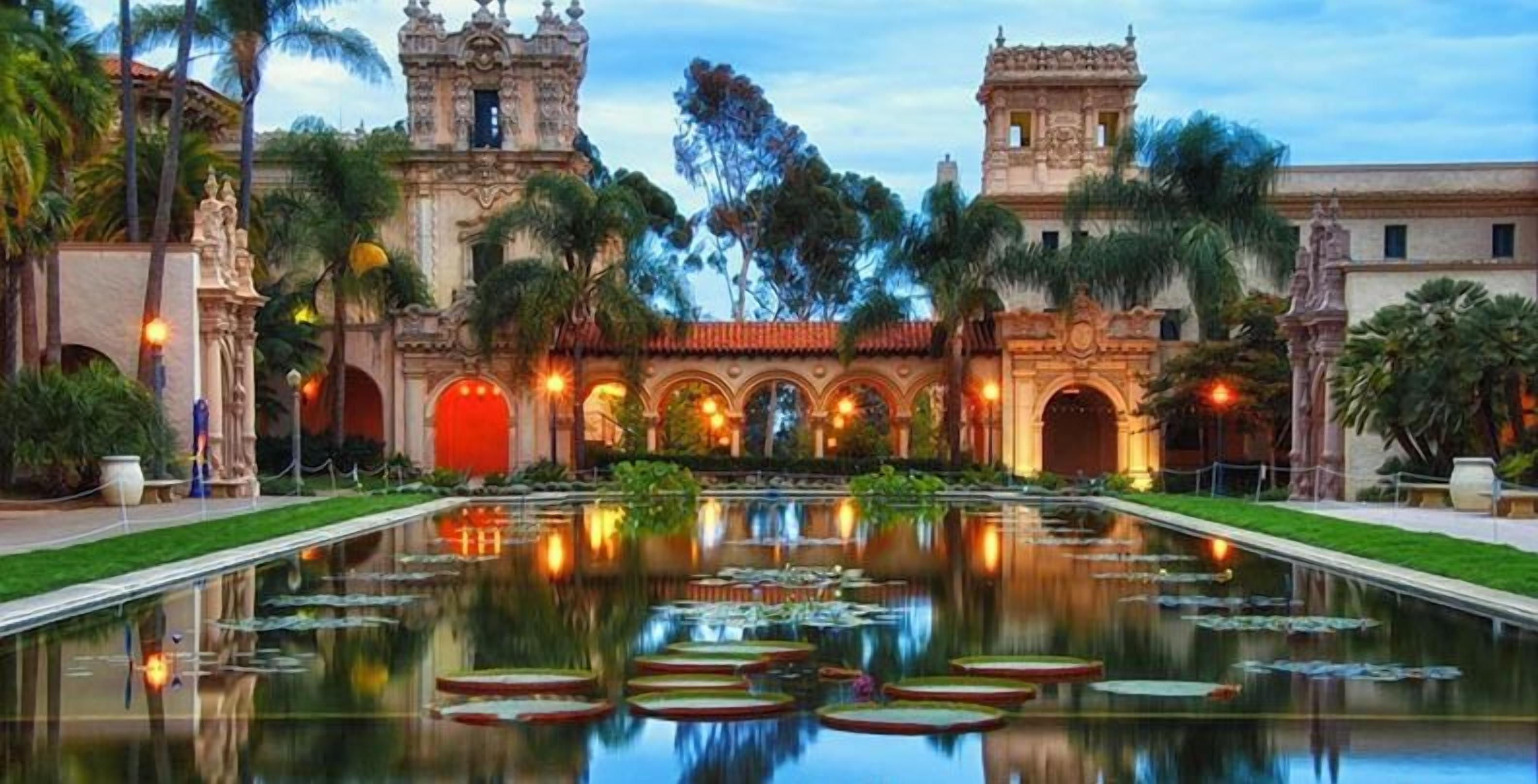 A courtyard with a reflective water feature, historic architecture, and palm trees.