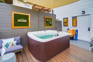 Un bain à remous dans un espace extérieur abrité, doté d’une terrasse en bois, d’un canapé avec des coussins à motifs et de deux éléments de décoration encadrés accrochés au mur.