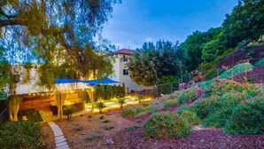 A modern house with a covered outdoor seating area, surrounded by landscaped gardens and a sloped terrain.