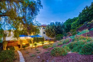 Una casa moderna con un espacio de estar cubierto al aire libre, rodeada de jardines ajardinados y un terreno inclinado.