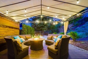 Un patio al aire libre con mobiliario de mimbre, una chimenea central y luces colgantes.