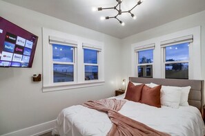 Un dormitorio con una cama, dos ventanas, un televisor en la pared y un moderno aplique de techo.