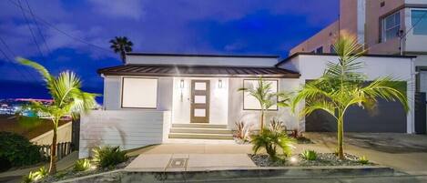 A modern two-story house with a white exterior, a front porch, and a small garden with palm trees.