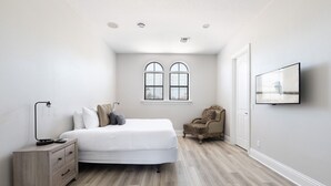 A bedroom with a bed, a chair, a nightstand, a TV, and a window with blinds.