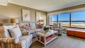 A living room with a view of the beach, featuring a striped sofa, a glass coffee table, and a red ottoman. - Oceanfront | Private Covered Balcony | Beautiful 4th Floor Amelia Surf and Racquet Condo (Fernandina Beach)