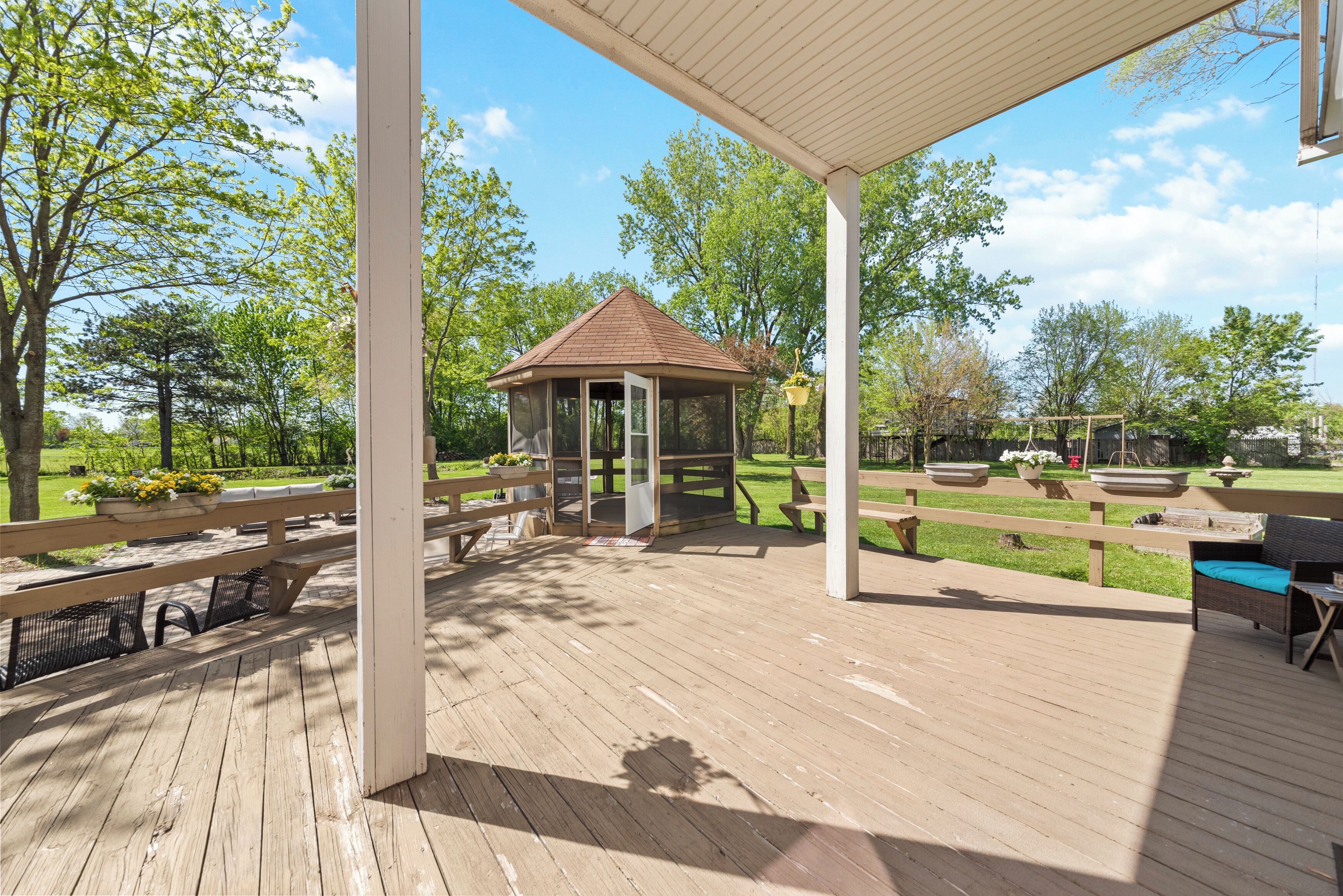 A wooden deck with a gazebo, picnic tables, and a view of a park with trees and a lawn.